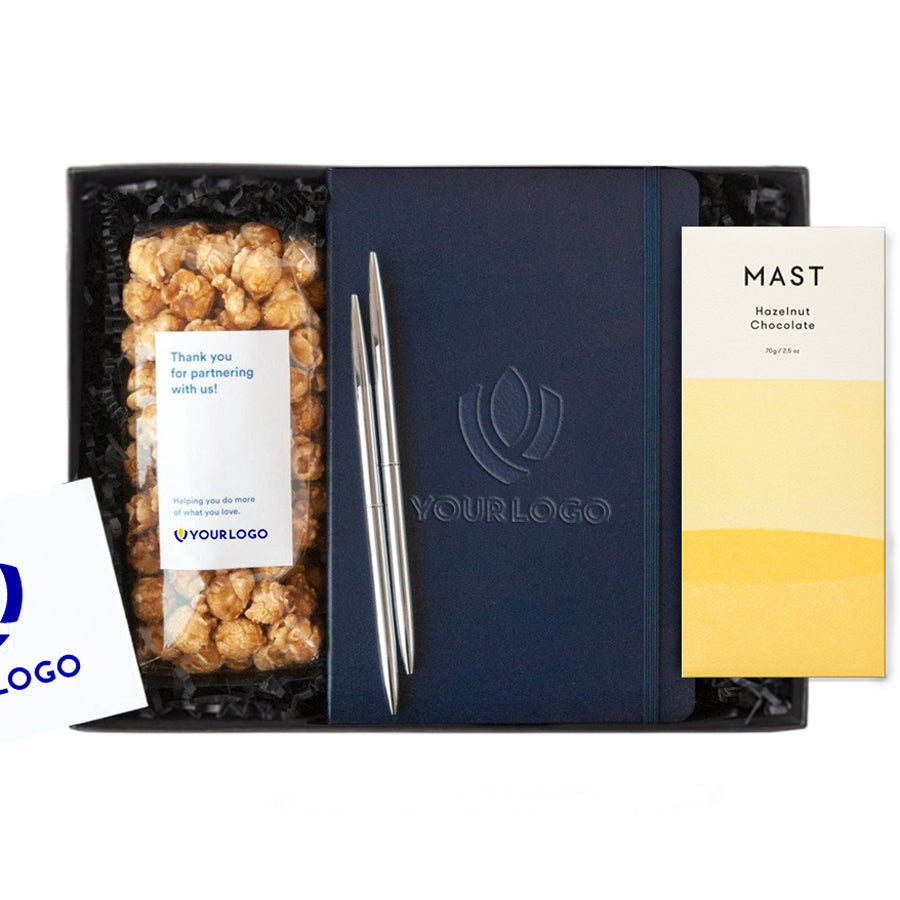 White client logo note book with blue text, set of two black pens, Large Mast oat milk chocolate with teal and white package, caramel chocolate drizzle popcorn with white and teal custom logo. All in white gifting box with white paper shred