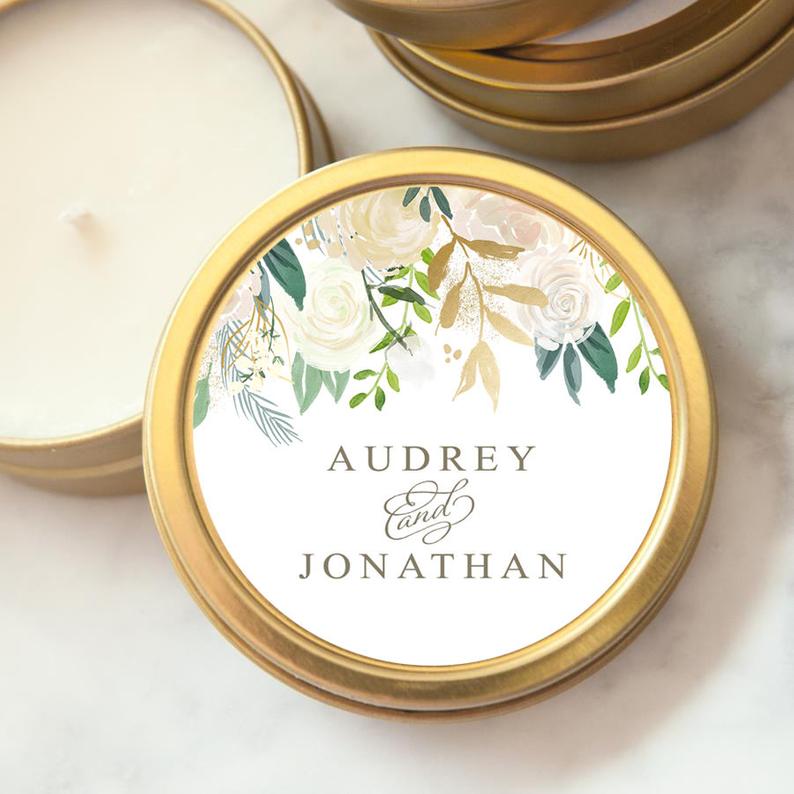 Close up image of Audrey custom wedding favor candle gold metal  container with a circular shape and a lid. On the lid, there is a white label that is decorated with floral arrangements.  The floral arrangement has white flowers and green leaves. Containers the name Audrey and Jonathan.  The candle is opened showing the white candle inside with another container on the upper right in a marble-like background.