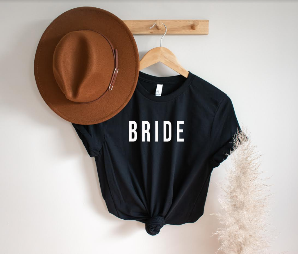 Black t-shirt on wooden hanger that reads "BRIDE" in white text. Between a brown hat and white plant.