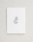 Our botanical card is featured. A minimalist, monochromatic sketch presents a sprig of delicate leaves centered on a stark white rectangular card, set against a neutral, textured backdrop.