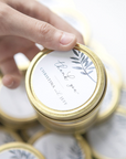 The image captures a close-up view of a hand holding a gold-colored, circular tin is a hand holding a gold-colored tin, likely made of metal. The top of the tin features a white label with the words "Thank you" in elegant script and "CHRISTINA and JEFF" underneath in smaller print. A watercolor-style sprig of leaves adds a decorative touch.