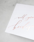 White envelope size card (approx. 3.5" x 5"), reads, "Will you be my bridesmaid?" in rose gold text zoomed in.