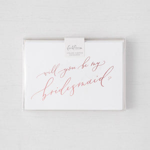 White envelope size card (approx. 3.5" x 5"), reads, "Will you be my bridesmaid?" in rose gold text in a clear plastic bag,