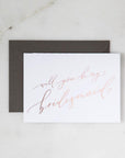 White envelope size card (approx. 3.5" x 5"), reads, "Will you be my bridesmaid?" in rose gold text. Slate gray envelope option behind.