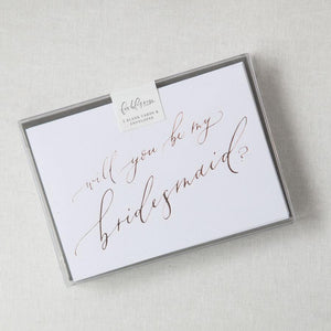 White envelope size card (approx. 3.5" x 5"), reads, "Will you be my bridesmaid?" in rose gold text in a clear plastic bag.