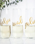 Three stemless flute glasses half half-filled with champagne gold script names (Chelsea, Nicole, and Julie). In a white background with a green branch on the background.