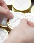 A close-up photograph displays a gold-colored candle tin featuring personalized details. a candle tin made of gold-colored metal, with a round shape and a visible seam. Inside the tin, there is a white candle. A circular label covers the top, displaying elegant calligraphy. The label features the names "Jenna & Mike," the hashtag "