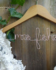 Light brown wooden hanger with a custom base wire shaped into the name 'Mrs. Fisher, with a bridal dress hanging.