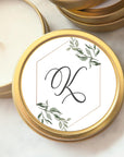 Custom wedding favor candle monogram, The candle is a small, round, gold-colored tin. On its top, there's a white label with a gold-colored hexagonal border. The label contains a stylized black cursive letter "K."and it is surrounded by delicate watercolor-style green foliage.