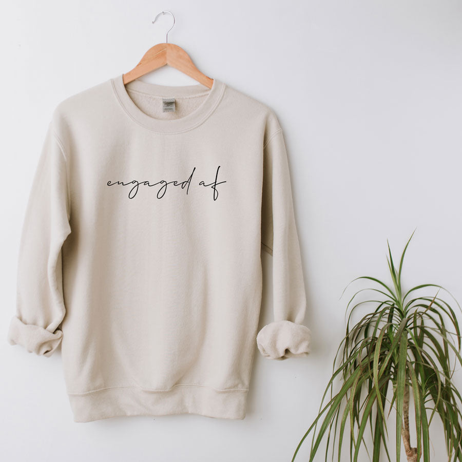 Custom Bride-to-Be Engagement or Bachelorette Sweatshirt with "engaged af" in lowercase script across chest, on white background with a green plant next to it.