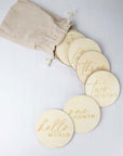 First year milestone discs presents a set of engraved wooden milestone discs for a baby, along with a small drawstring bag, arranged on a neutral, textured surface. The discs are arranged in a slight arc, creating a visually appealing composition that guides the viewer's eye. The words “hello world”, “one month”, “two months”, “three”, “four” and "six months" are visible, along with others partially visible.