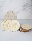 The image showcases a collection of wooden milestone discs for a baby, along with a small canvas bag, arranged on a light-colored surface,arranged on a solid, light-colored surface, likely a tabletop, with a light-colored marble or stone backdrop.