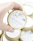 The image showcases a hand holding a gold-rimmed container with a personalized label for a wedding, surrounded by several similar containers,It's made of gold metal with a circular shape and a lid. On the lid, there is a white label that is decorated with floral arrangements. The label contains the names "Jen" and "Vik" in a stylish script font, beneath which the date "July 14, 2018" is printed. The floral arrangement has white flowers and green leaves.