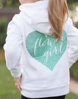 The image shows the back of a girl wearing a white hoodie featuring a jade-colored heart with the words "Flower Girl" in script cut out from it in a nature background.