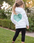 The image shows the back of a girl wearing a white hoodie, black leggings, and tennis shoes, featuring a jade-colored heart with the words "Flower Girl" in script cut out from it against a natural background.