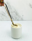 Gold wick trimmer being held by a hand trimming a candle in a white jar.