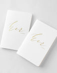 Her and her vow books, white linen cover, metallic gold foil stamping, rounded corners, saddle stitch binding featured on a white background.