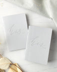 His and hers gray linen letterpressed vow books, featured in a marble-like background with flowers on one side and a linen cloth on the other.