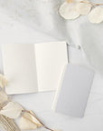 His and hers gray linen letterpressed vow books, one is opened and the other showing the back detail, featured in a marble-like background with flowers on one side and a linen cloth on the other.