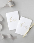 His and hers white, rounded-corner, gold-foil-stamped vow books featured in a gray background, with a gold pen on the right side.