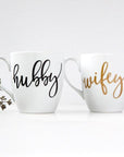 White ceramic mugs, two 15 oz mugs, "hubby" in black script font, "wifey" in gold script font.