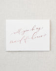 White envelope size card (approx. 3.5" x 5"), reads, "Will you be my maid of honor?" in rose gold text.