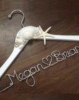 White hanger with shells and starfish accents. Personalized handcrafted wire hanger base in silver aluminum personalized with Megan, heart shape, and Brian on a dark wood background.