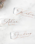 Personalized bridal party stemless flutes, featuring three transparent designs with rose gold lettering reading from top to bottom: Katie – Maid of Honor, Alex – Bride, and Lindsay – Bridesmaid, set against a marble-like background with flowers in the top left corner.