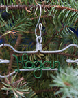 Personalized Dog Ornaments, hanging in a Christmas tree as a background, are made of silver-colored aluminum in a dog bone shape outline with the name 'Hogan' in green-colored wire.
