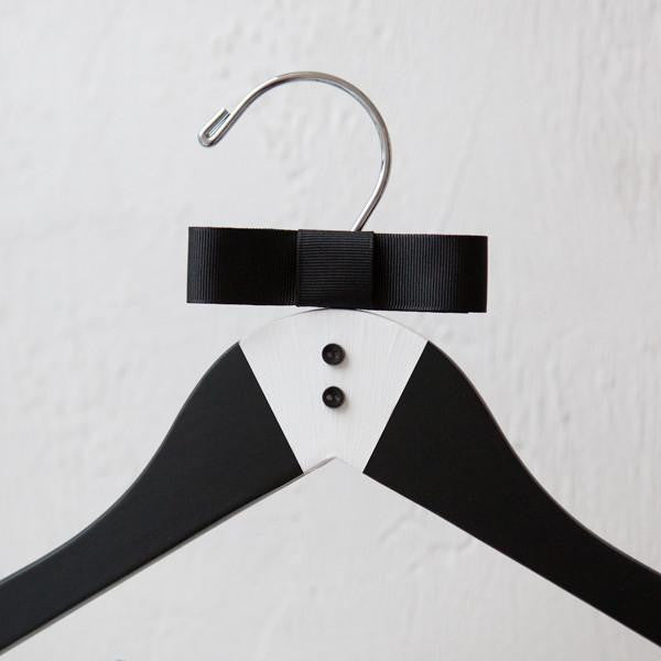 Close up of luxe matte black hanger, hand-painted with a tuxedo design and finished with two tiny buttons and a grosgrain bowtie with silver hock. Ona white background.
