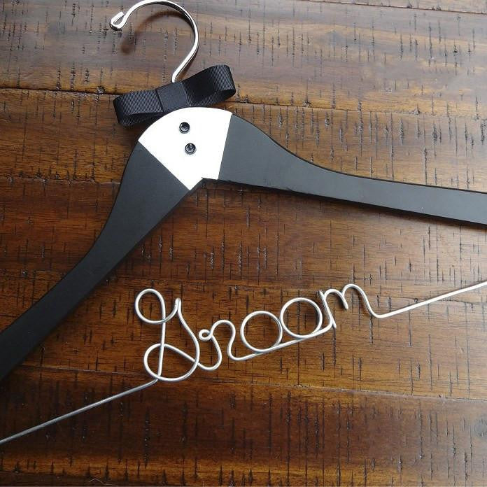 A luxe matte black hanger, hand-painted with a tuxedo design and finished with two tiny buttons and a grosgrain bowtie, customized with "Groom," in silver wire with notches. On a wooden background.