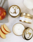 Featuring three gold tin jar candles labeled “Spiced Apple Cider.” The candles are displayed in different positions: one viewed from the top with its lid on, one placed sideways to reveal the side label, and one opened to show the smoot white wax inside. Each tin has a clean white label with elegant gold lettering and a delicate gold illustration of an apple tree on the lid. Set against a soft background with scattered red apple slices and white rose petals. A folded grey cloth in the corner.