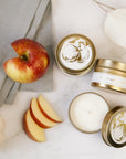 Featuring three gold tin jar candles labeled “Spiced Apple Cider.” The candles are displayed in different positions: one viewed from the top with its lid on, one placed sideways to reveal the side label, and one opened to show the smoot white wax inside. Each tin has a clean white label with elegant gold lettering and a delicate gold illustration of an apple tree on the lid. Set against a soft background with scattered red apple slices and white rose petals. A folded grey cloth in the corner.