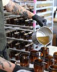 A teakwood and tobacco candle is shown being poured into its jar by a man with black gloves in the production headquarters.