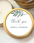 Close-up image of the thank-you custom wedding favor candle in its gold-rimmed tin, each with a label reading "Thank You" along with the names "Jessica and Jordan." A watercolor-style depiction of a leafy branch in shades of grey-blue is placed near the top. The lid opened, showing the white candle on a marble-like background.