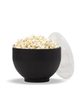 The Popper, a lack silicone popper expanded into a bowl holding popcorn with a clear plastic lid propped against the side, featured on a white background.