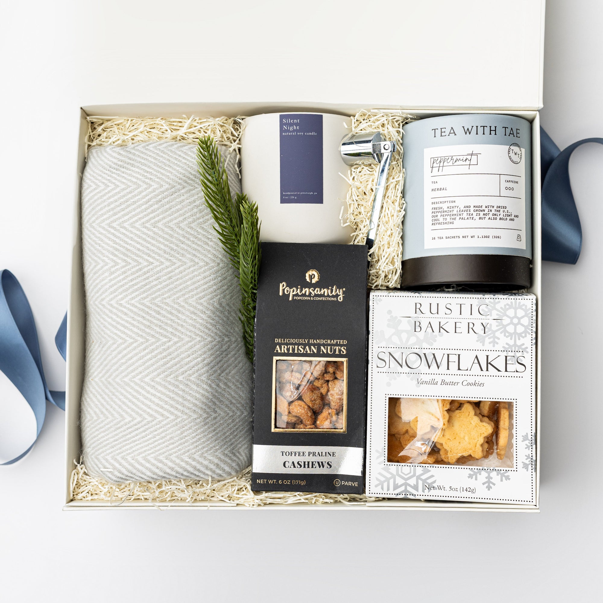 top down photo of a holiday themed cream gift box featuring a light gray herringbone throw blanket, a white ceramic candle with blue label named Silent Night, metal silver candle snuffer, grey canister of peppermint tea sachets, black with silver foil detail box of artisanal cashews, white box of shortbread cookies snowflake shaped with silver foil snowflakes print, preserved spruce leaf ornament. All items are nicely nested on ivory thin shredded paper with winter-blue ribbon around the box.