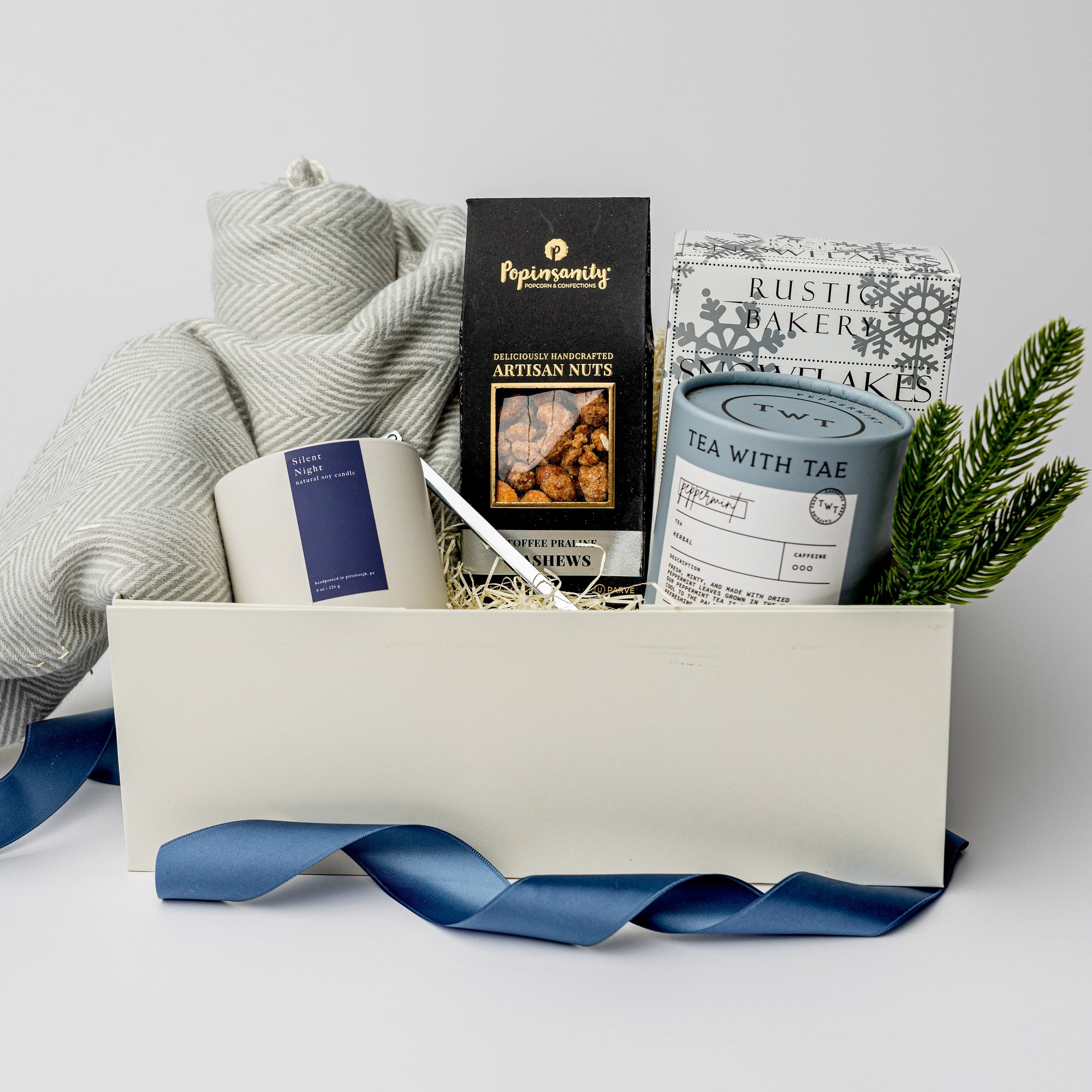 top down photo of a holiday themed cream gift box featuring a light gray herringbone throw blanket, a white ceramic candle with blue label named Silent Night, metal silver candle snuffer, grey canister of peppermint tea sachets, black with silver foil detail box of artisanal cashews, white box of shortbread cookies snowflake shaped with silver foil snowflakes print, preserved spruce leaf ornament. All items are standing, staged inside the box with winter-blue ribbon around the box.