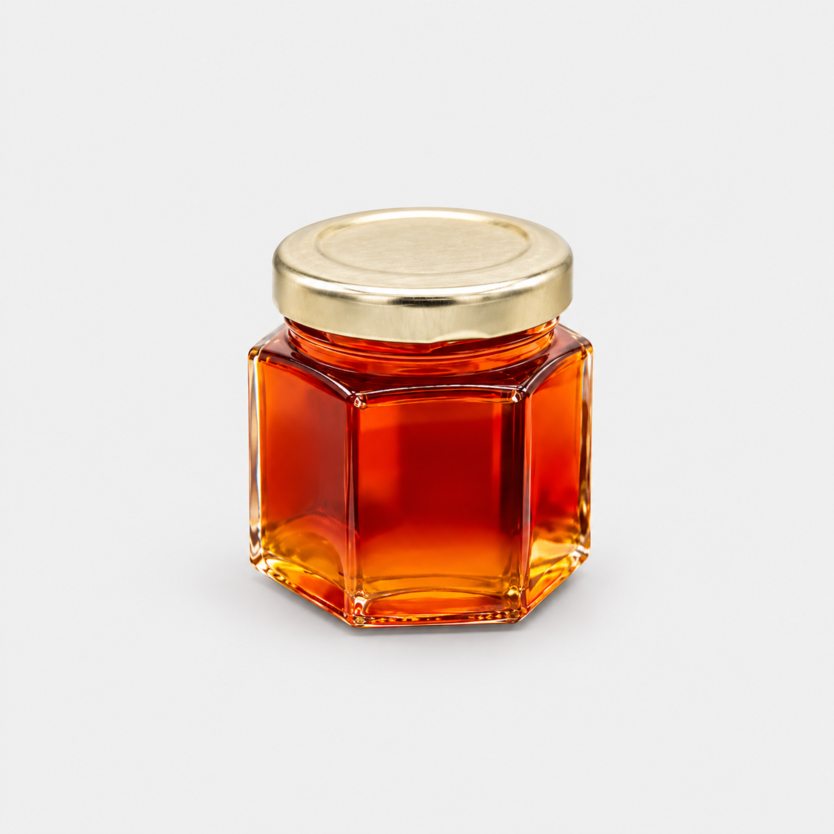 A small hexagonal glass jar filled with amber-colored honey, featuring a shiny gold metal lid, set against a plain light gray background.
