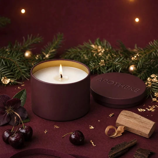 A lit candle in a matte burgundy cylindrical tin with a gold interior, shown beside its matching lid engraved with the brand name 'APOTHEKE'. The scene is styled with dark cherries, pine branches, gold leaf, and wood pieces on a deep burgundy surface against a dark burgundy background.