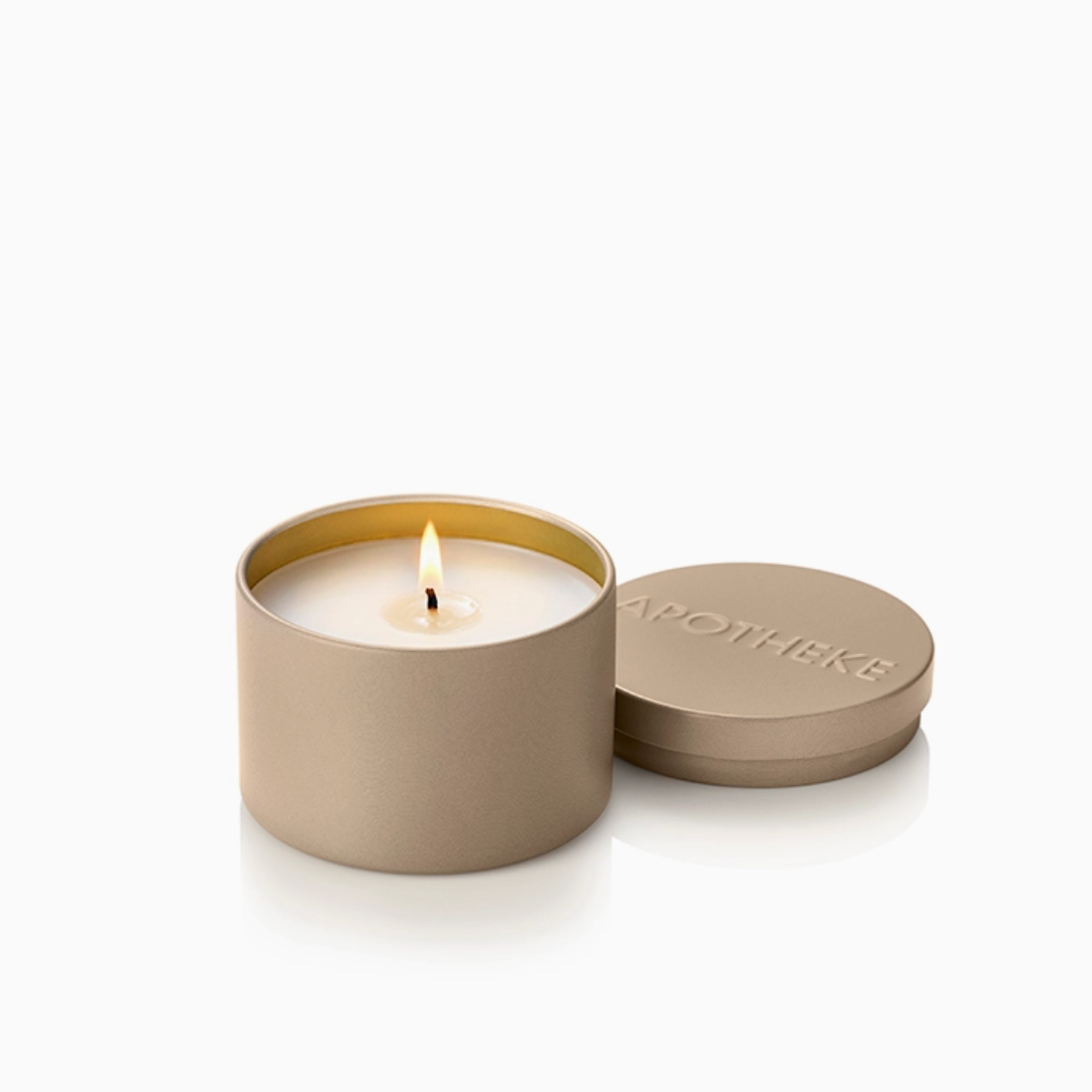A lit candle in a matte taupe metal tin with its lid resting beside it on a clean white background. The embossed brand name 'APOTHEKE' is visible on the lid.