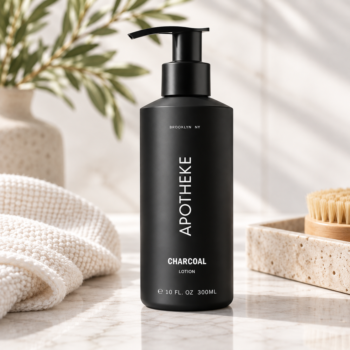 A black pump bottle of APOTHEKE Charcoal Lotion stands on a light marble surface with a folded cream towel, a wooden brush, and soft plant shadows in the background.
