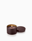 A lit cylindrical candle in a dark brown matte tin with its lid beside it, embossed with the brand name 'apotheki'.