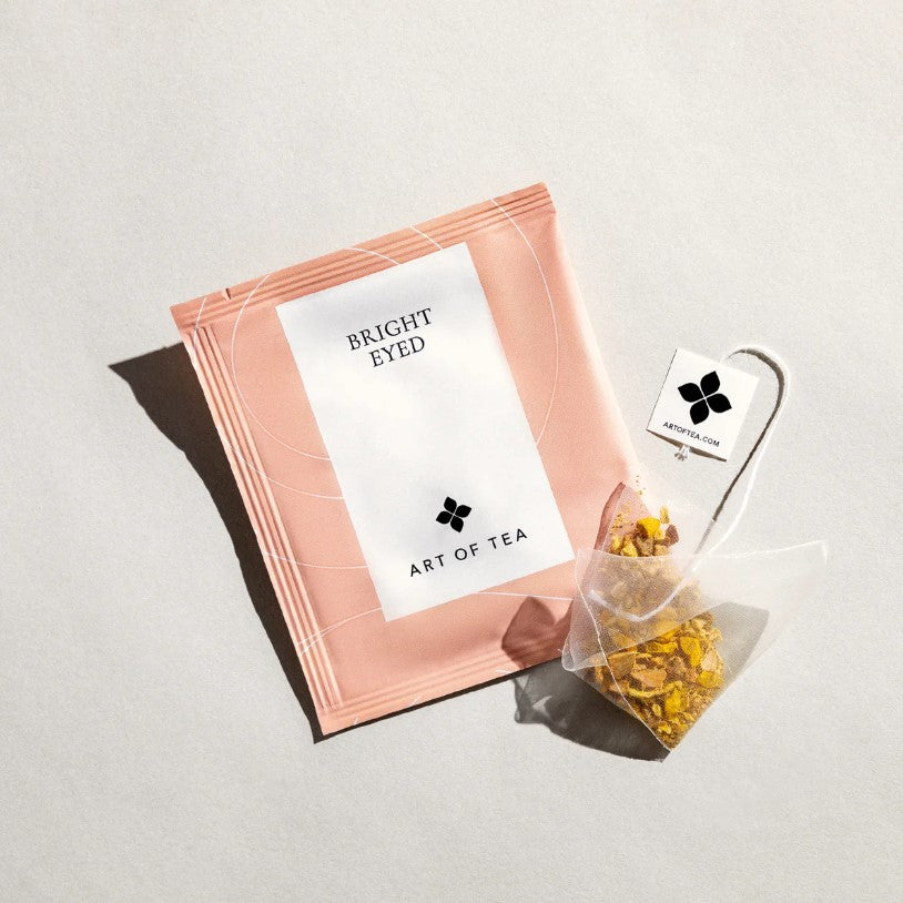 Bright eyed tea from art of tea is featured in its pink square shaped with white label and black lettering and next to it the tea outside of its sachet package on a white background. 