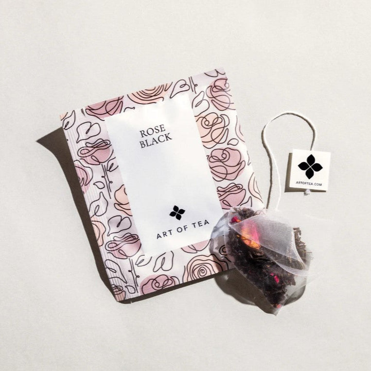 Art of tea rose black tea squared-shaped sachet  package is featured in its pink roses illustration with the label in white in black lettering and the tea outside the tea sachet package next to it ion a white background.  