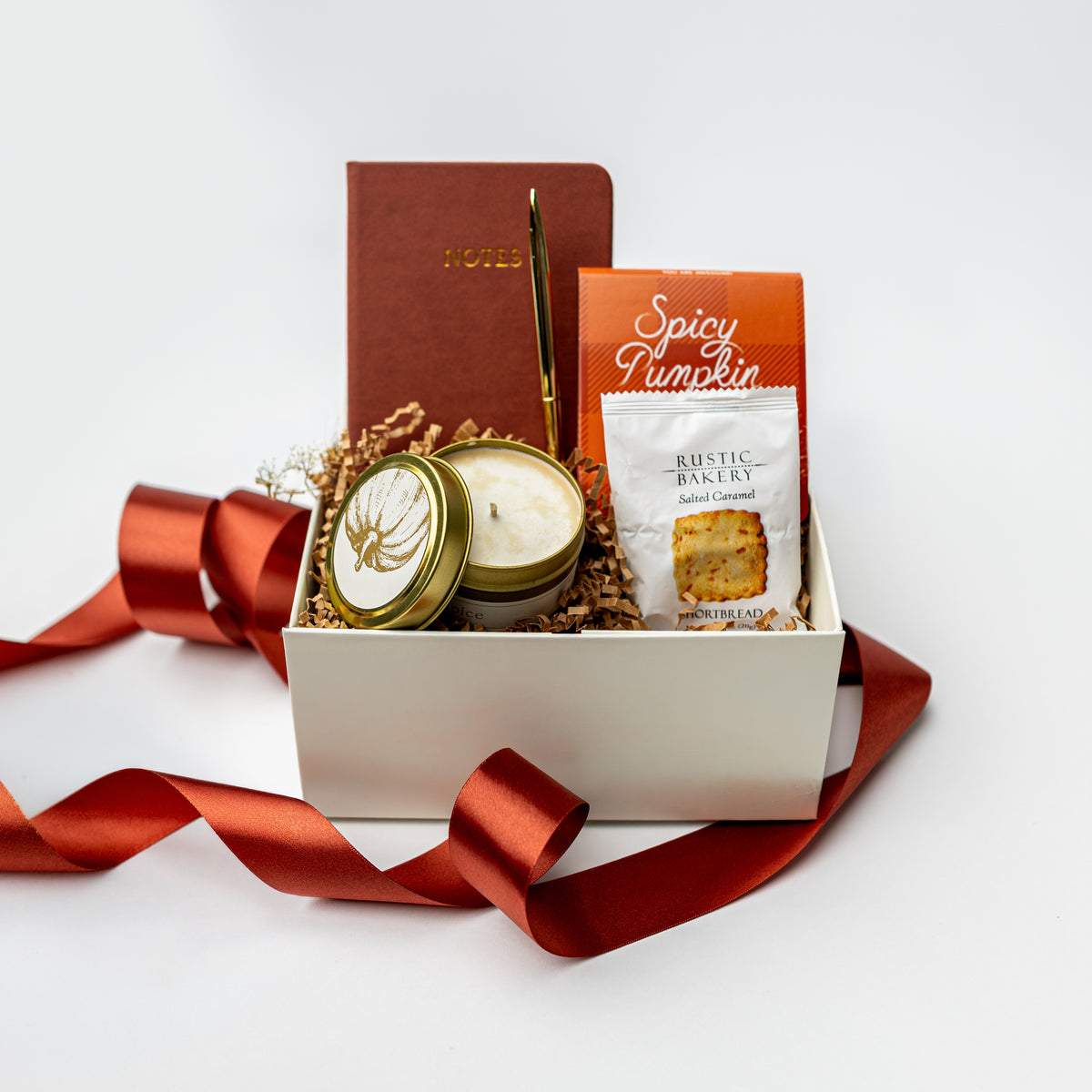 A curated gift box set on a white background, featuring a white box filled with a brown notebook, a candle tin, a candle, a bag of Rustic Bakery Spicy Pumpkin cookies, and a gold pen, with a red ribbon draped around it.