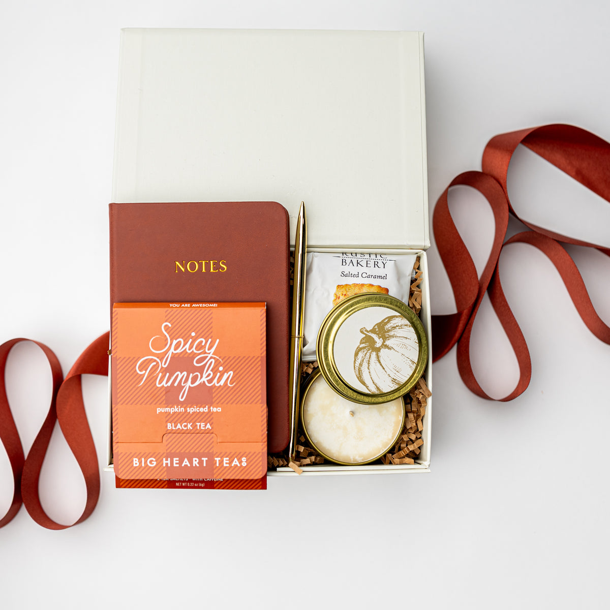 A curated gift box set on a white background, featuring a brown notebook, a box of Big Heart Teas Spicy Pumpkin black tea, a small round tin of baked cream, and a gold pen, arranged with a rust-colored ribbon.