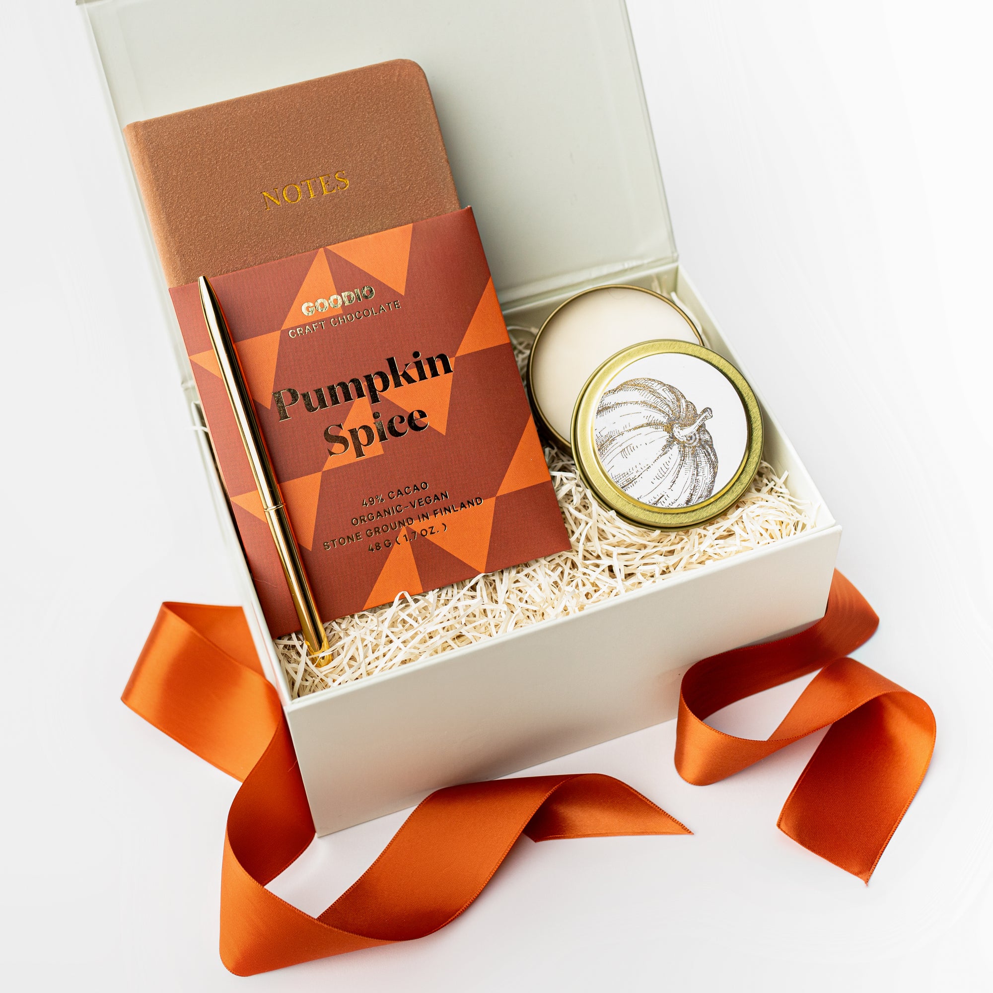 Fall Autumn Seasonal gift box containing pumpkin spice chocolate, pumpkin spice candle, gold pen, and small linen taupe notebook inside of a white box with ivory shred, and ornage ribbon. 
