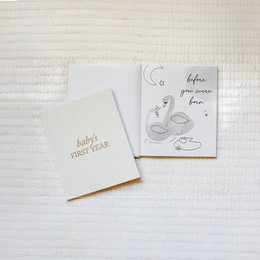 Two greeting cards on a textured white surface, one with a swan illustration and text, the other with 'baby's first year' text.