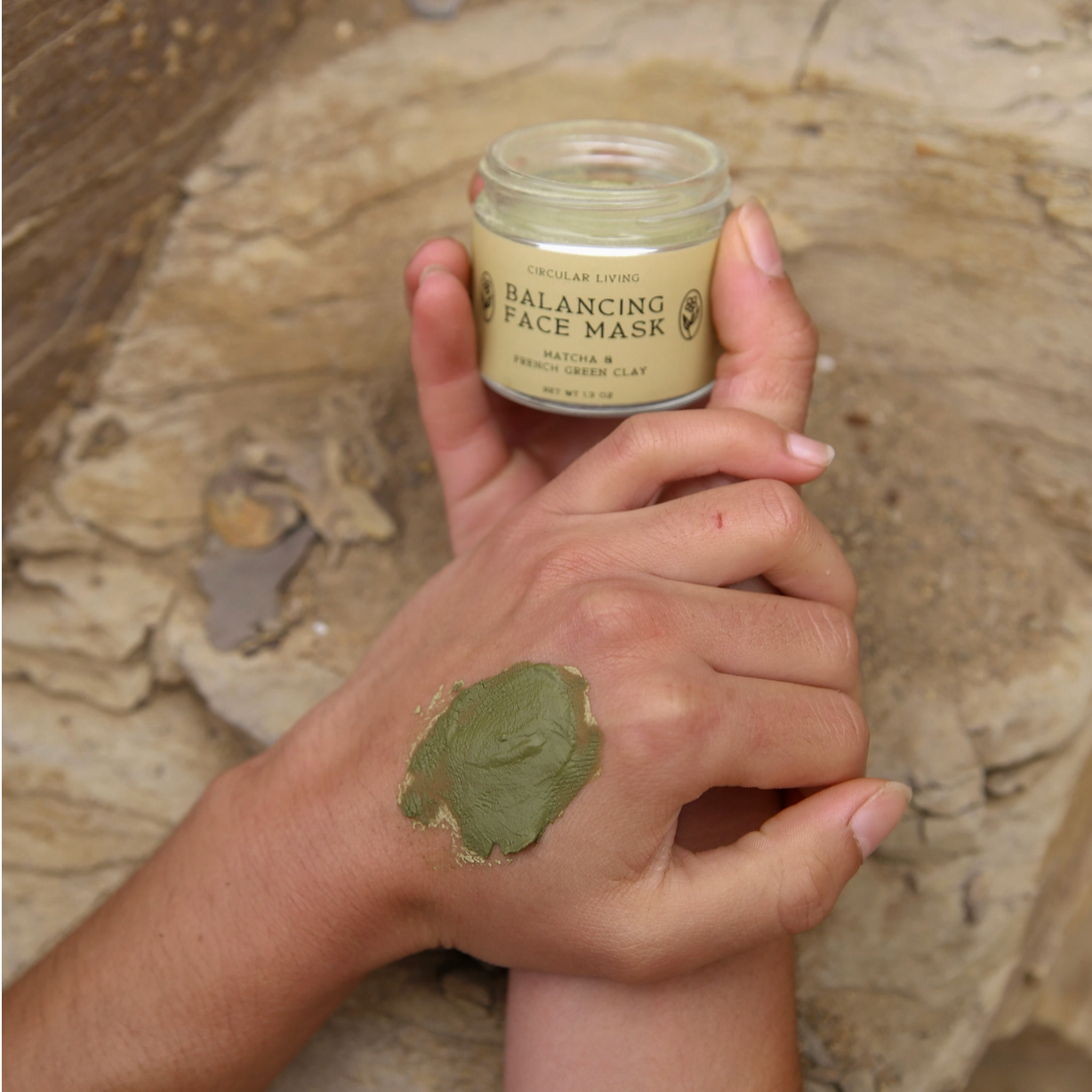 Hand holding a jar of Balancing Face Mask with green mask on wrist against a stone background